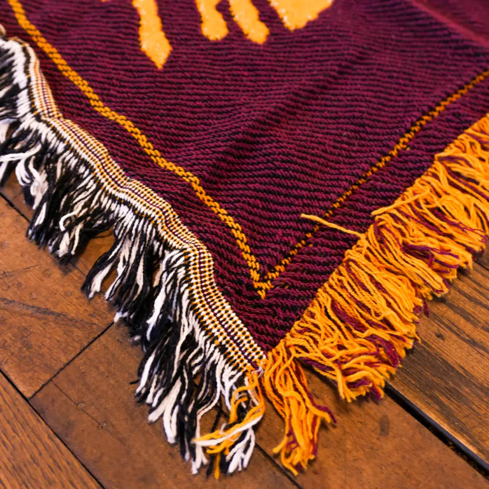 The Revivalists woven blanket features a rich maroon woven blanket with bold orange geometric patterns and black and white fringe