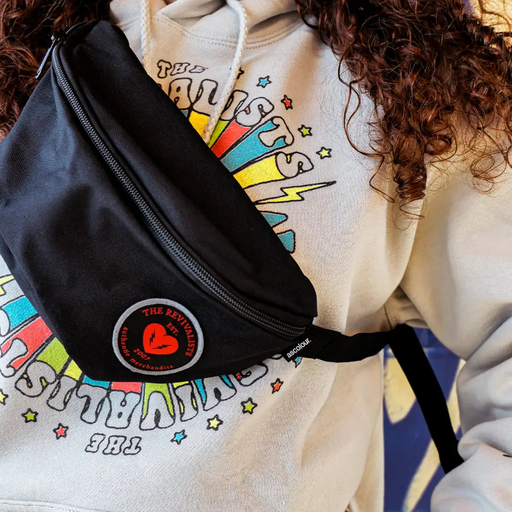 Black fanny pack with a red and white circular logo worn over a colorful graphic sweatshirt.